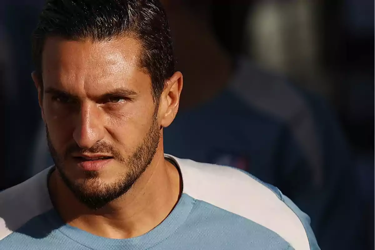 Man with a beard and dark hair, wearing a light blue t-shirt, looking forward with a serious expression.