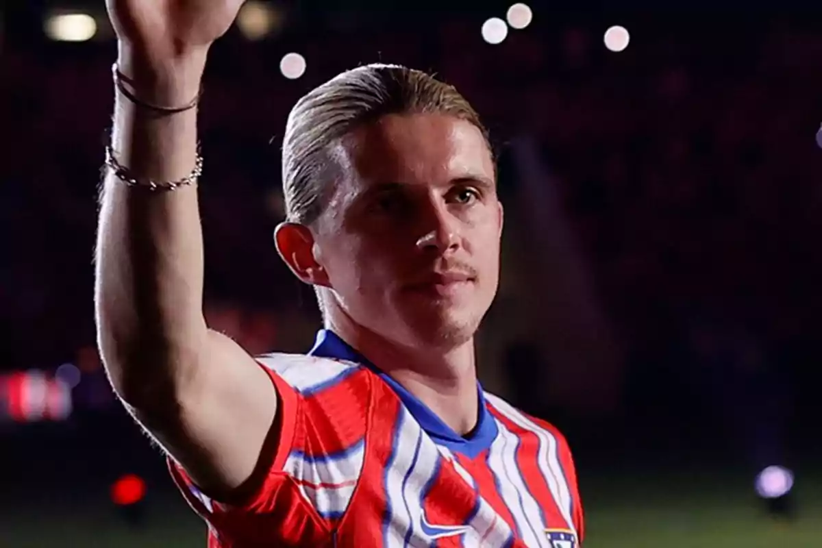 A soccer player in a red and white uniform raises his hand in a floodlit stadium.