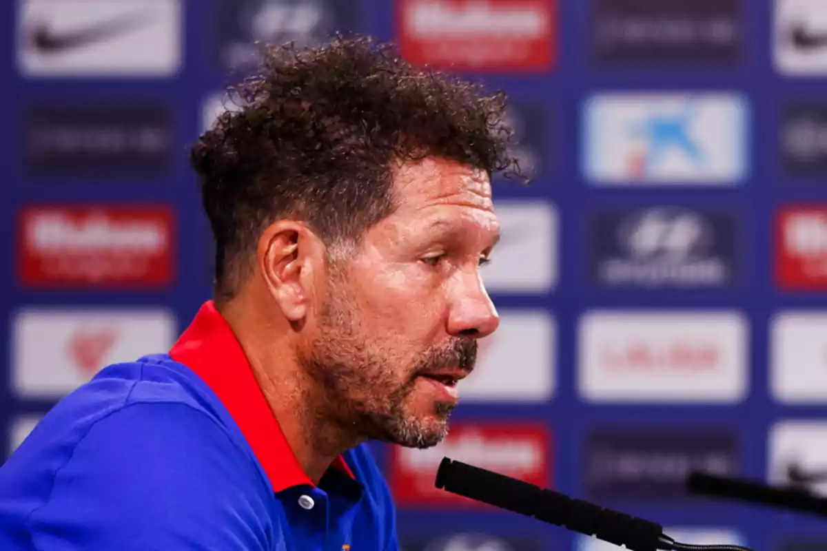 A bearded man with curly hair speaks at a press conference, against a background of sponsor logos.