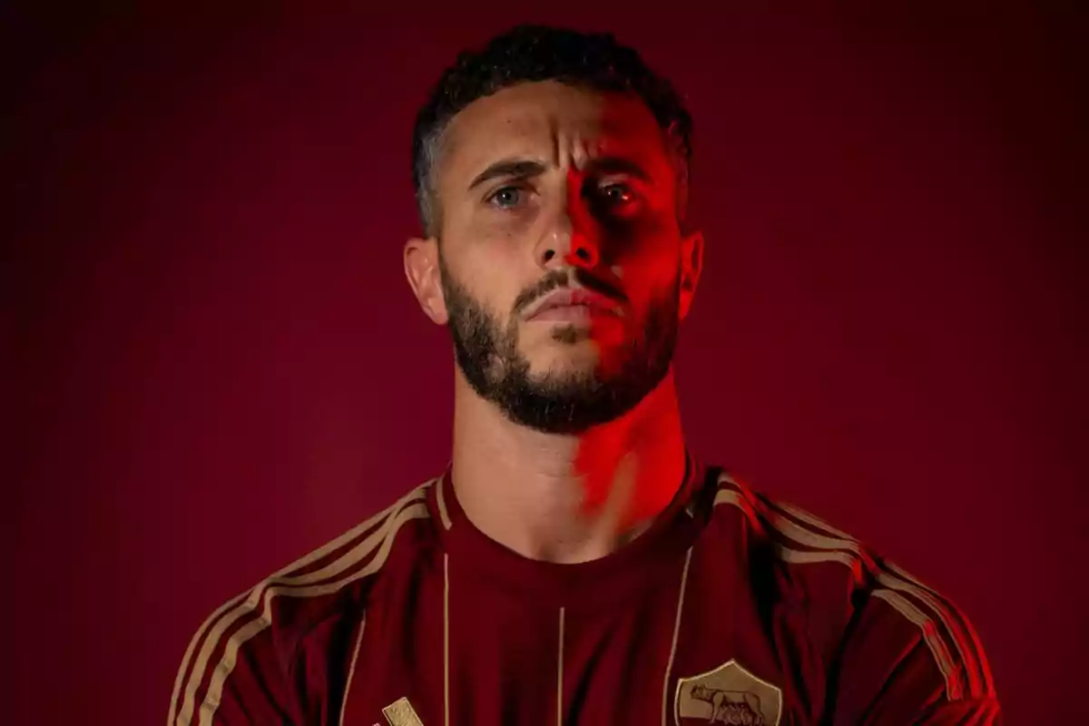 Man with beard and curly hair, wearing a red sports shirt with gold details, illuminated with red light on a dark background.