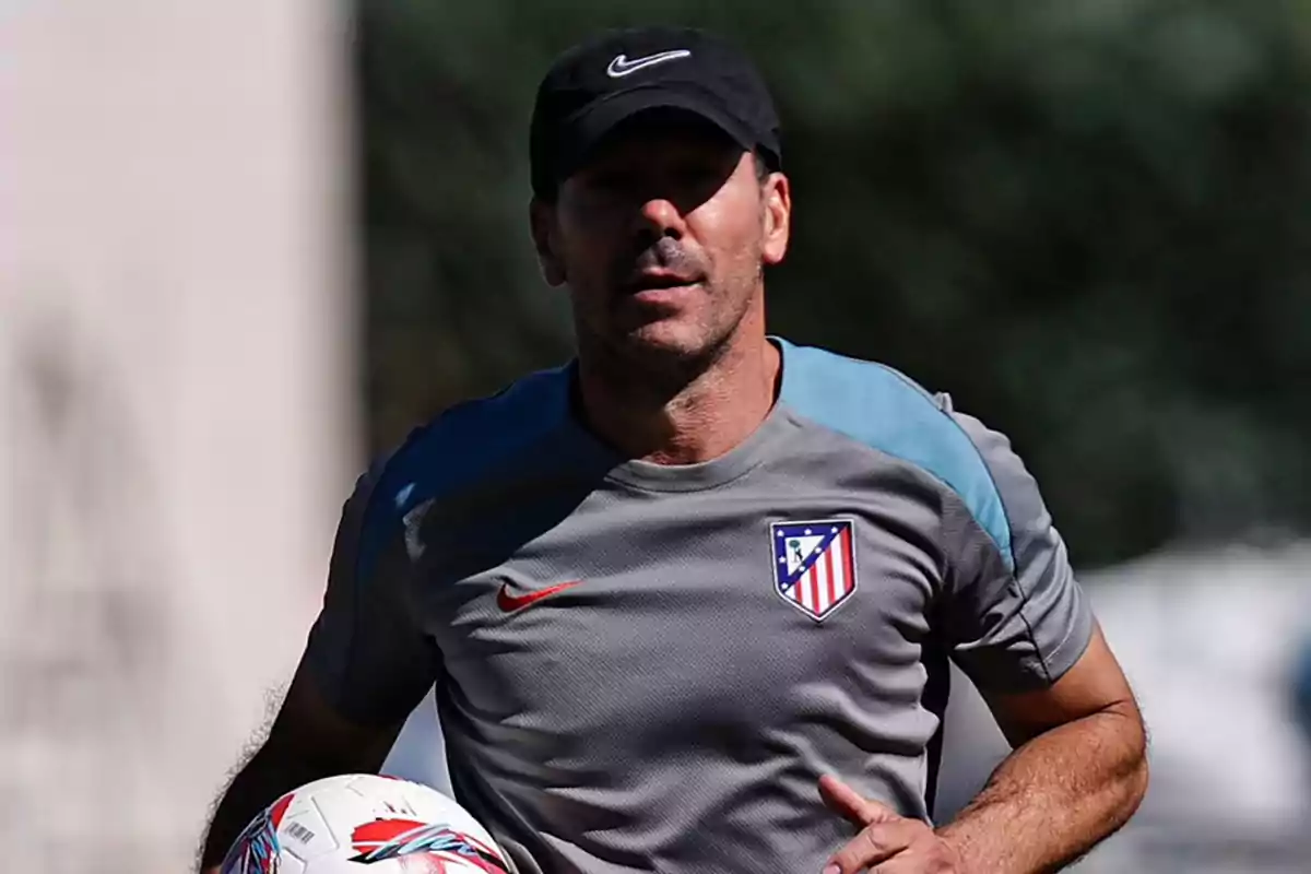 A man wearing a black cap and a grey Atlético de Madrid shirt holding a football.