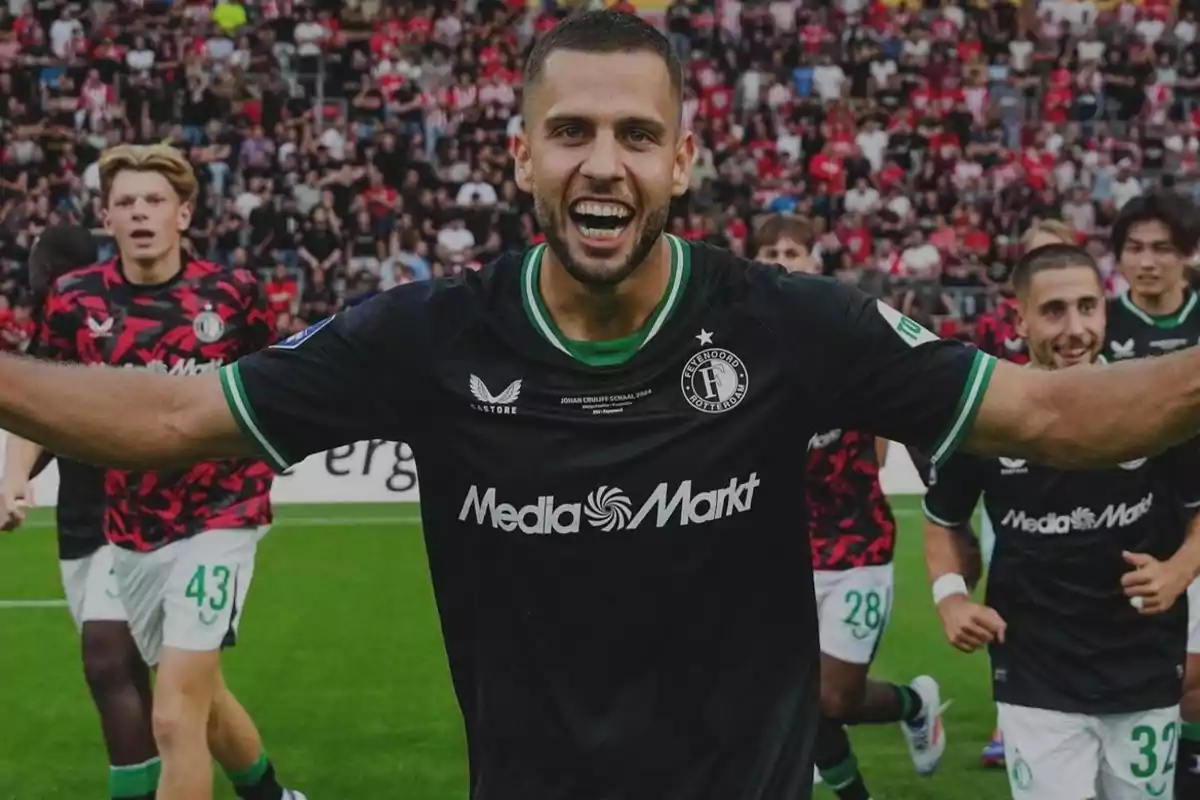 A group of soccer players celebrating on the field with one player in the foreground smiling and spreading his arms.