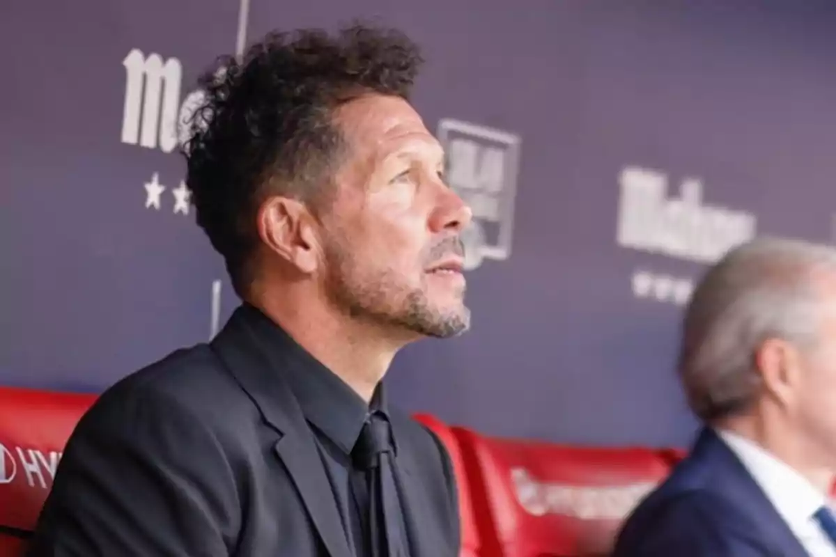 A man in a black suit with a beard looks up while sitting on a red bench, with a background showing logos and blurred text.