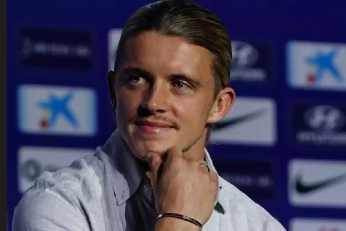 A man in a light shirt and with his hair tied back poses with his hand on his chin in front of a blue background with logos.