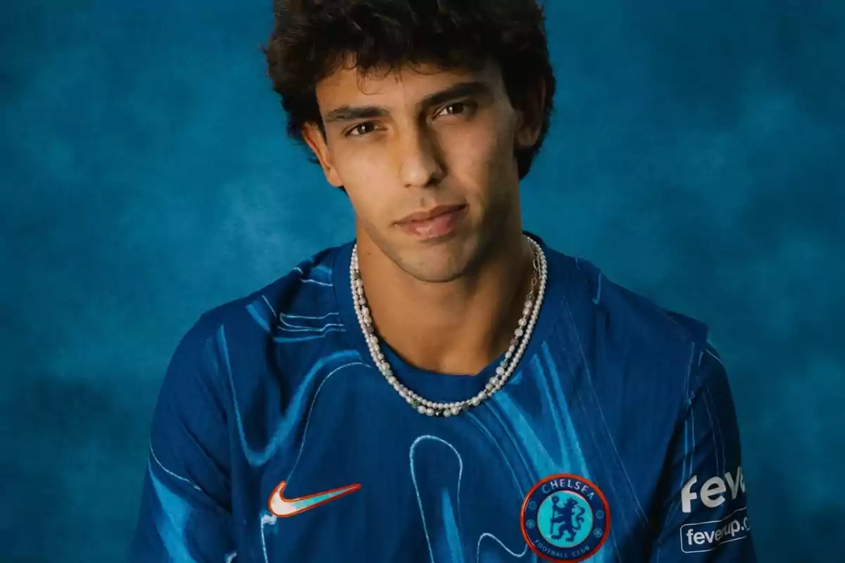 A man wearing a blue Chelsea FC shirt and a pearl necklace, posing in front of a blue background.