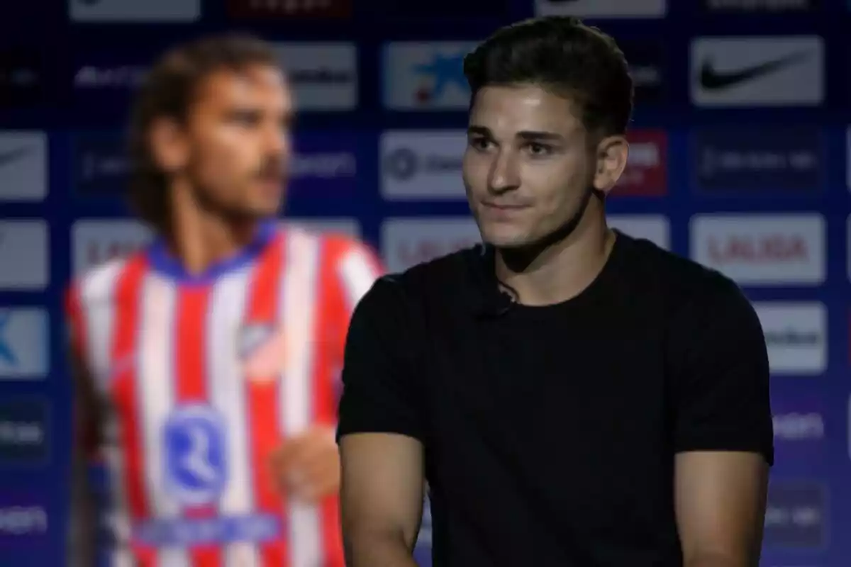 A man in a black T-shirt sits in front of a blurred background showing a football player in a red and white striped shirt.