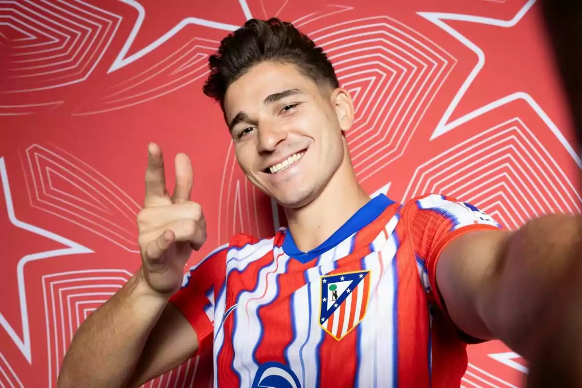 A soccer player wearing an Atletico Madrid shirt smiles and gestures with his hand as he takes a selfie in front of a red background with geometric patterns.