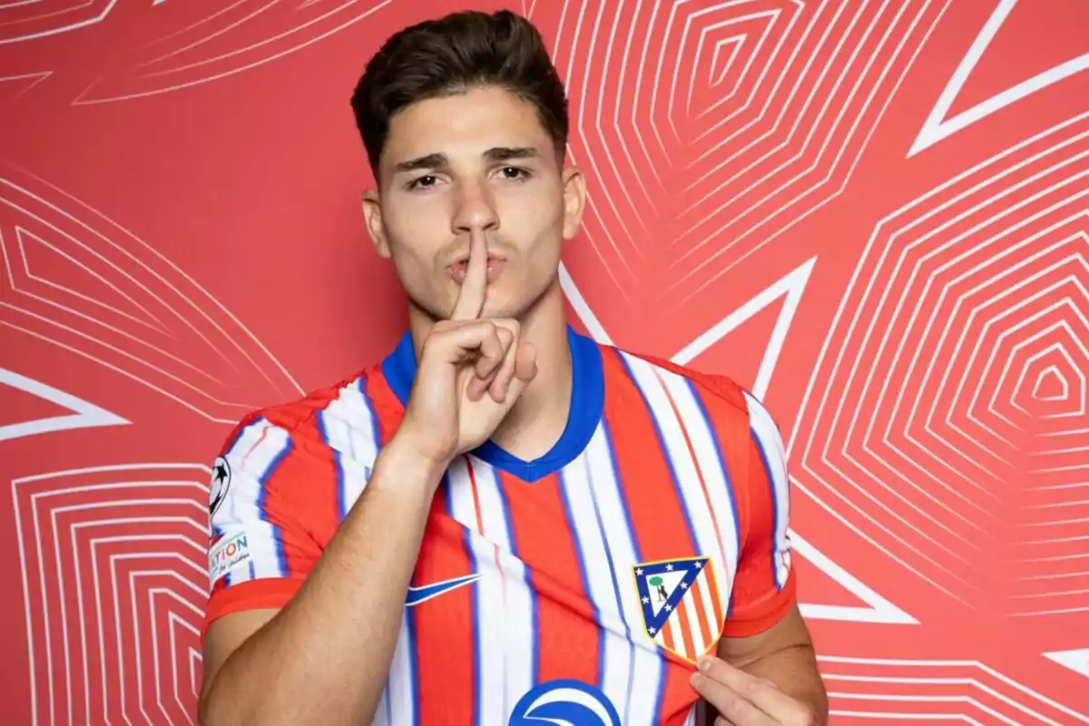 A football player wearing an Atlético de Madrid shirt makes a silence gesture with his index finger on his lips in front of a red background with geometric patterns.