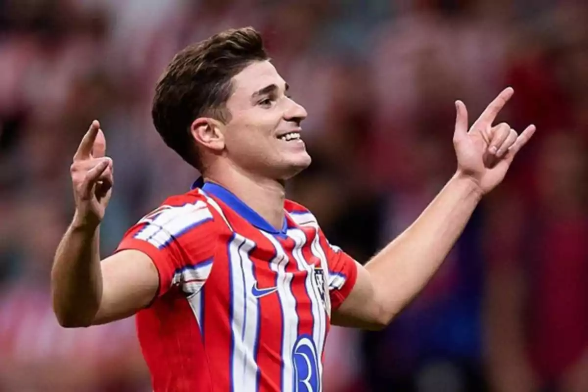 A soccer player celebrating a goal with his arms raised and smiling, wearing a red, white and blue striped jersey.