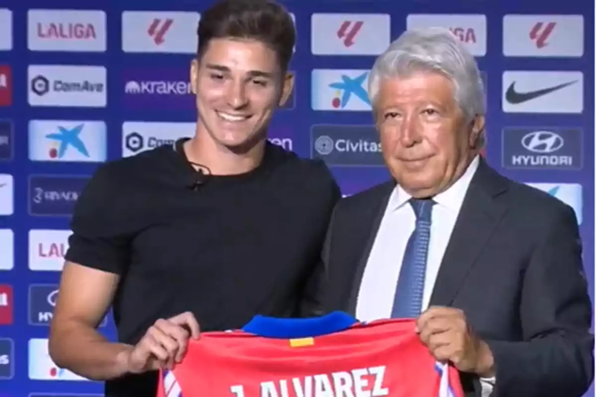 Two people pose for a photo holding a football shirt with the name "J. Alvarez" on it at an event with various sponsor logos in the background.