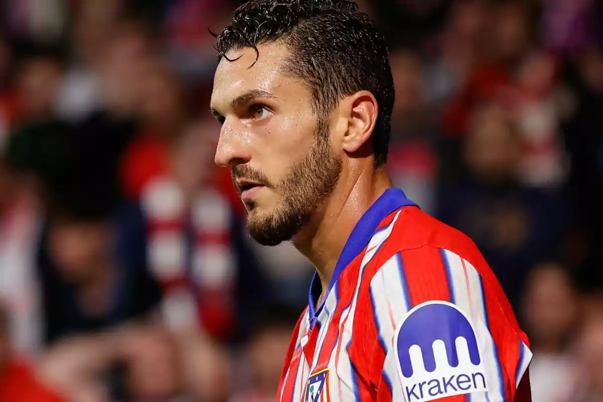 A soccer player wearing a red and white striped jersey with the Kraken logo on the sleeve stands on the field, sweating and looking to the right.