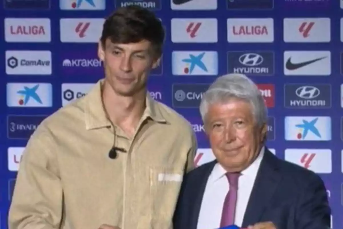 Two men pose together in front of a background with logos of sports sponsors.