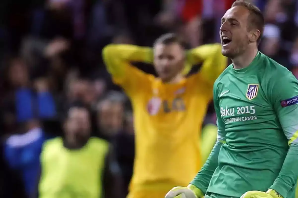 A soccer goalkeeper in a green uniform celebrates while another player in a yellow uniform looks disappointed in the background.