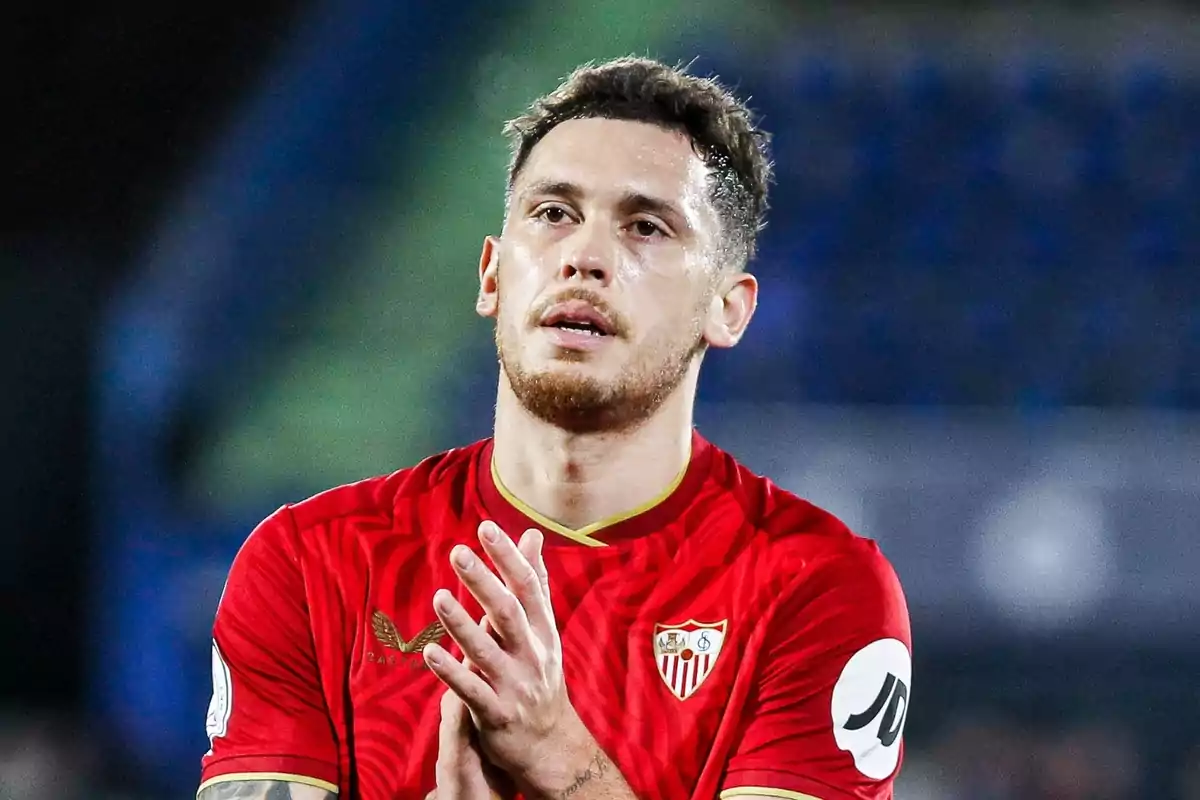 A football player wearing the red Sevilla FC shirt applauding on the pitch.