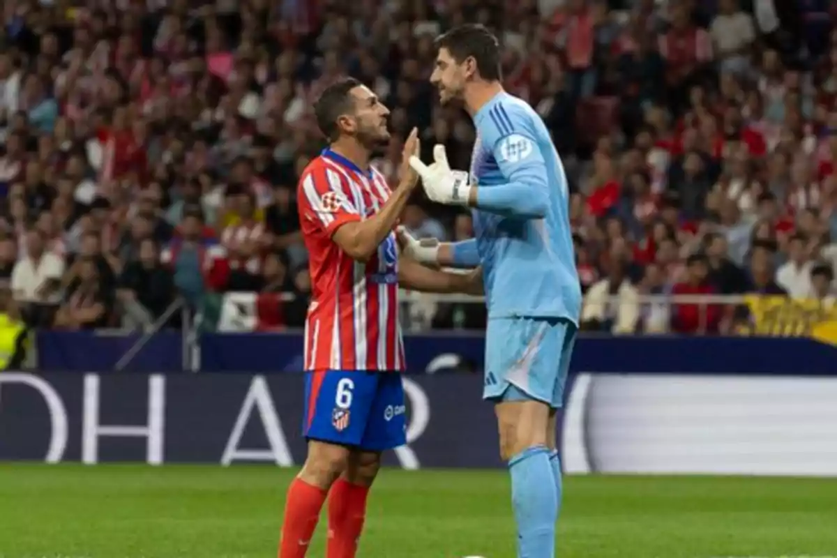 Two soccer players argue on the field during a match, one in a red and white uniform and the other in a blue uniform.