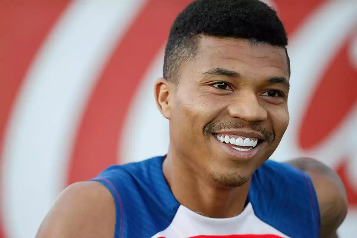 A smiling man wearing a blue and white sports t-shirt, with a red and white out of focus background.