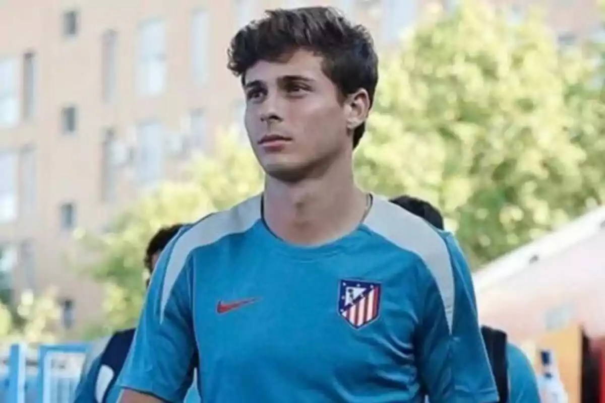 A young man in a blue Atlético de Madrid shirt is standing outdoors with a building and trees in the background.