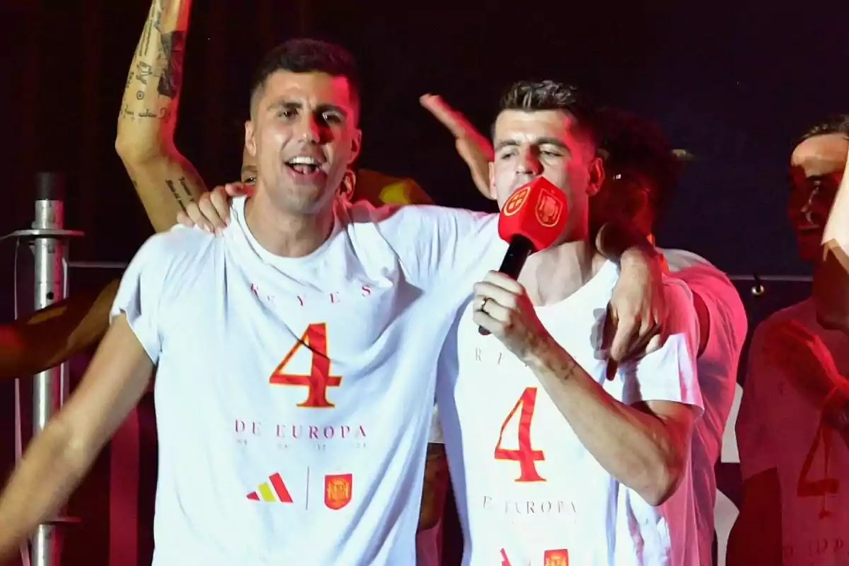 Rodri and Álvaro Morata celebrate with white t-shirts that have the number 4 and the phrase "Kings of Europe", Álvaro holds a red microphone.