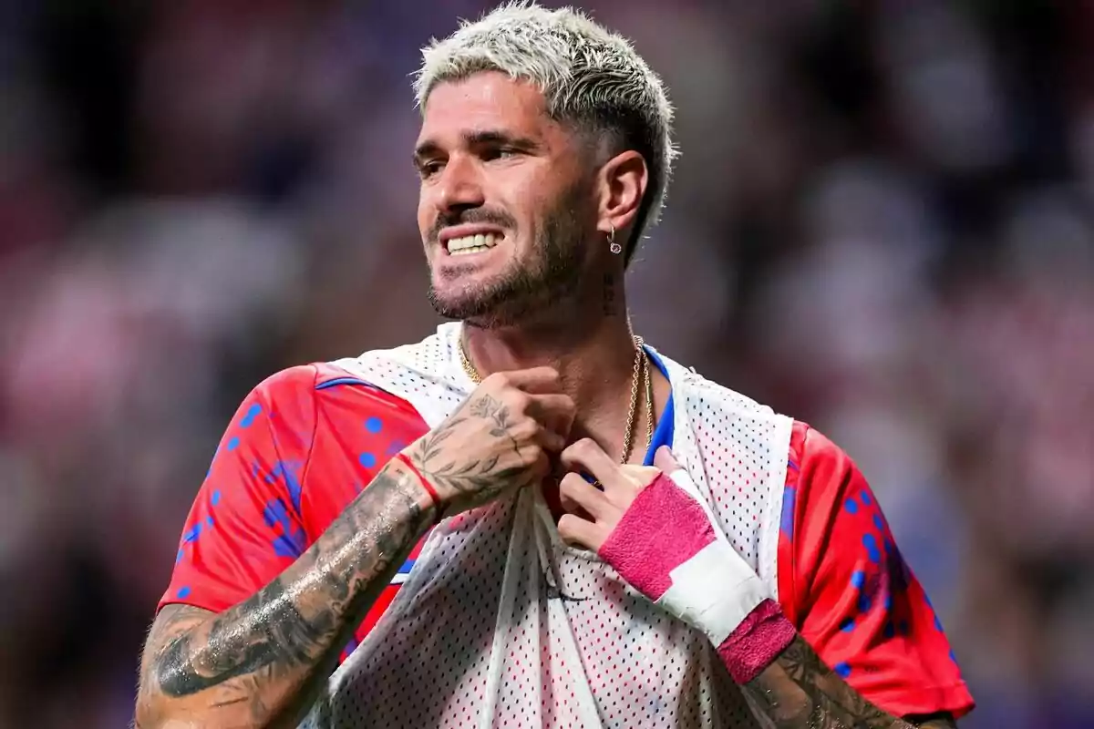 A soccer player with blond hair and tattoos on his arms, wearing a red and blue jersey and a pink bandage on his wrist, smiles as he adjusts a white bib.