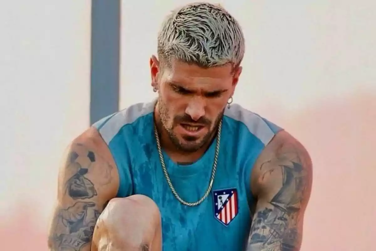 A man with platinum blonde hair and tattoos on his arms, wearing a blue sleeveless T-shirt with the Atlético de Madrid crest, appears to be concentrating or tired.