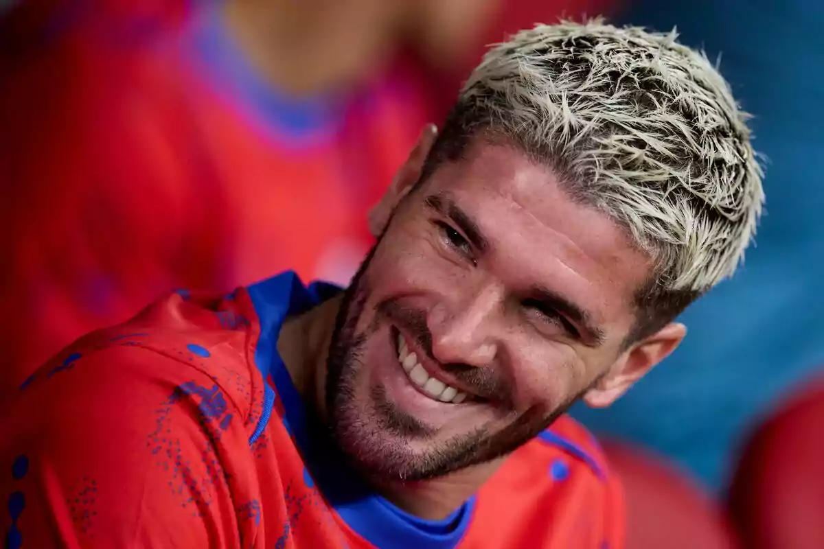 A man with platinum blonde hair and a beard smiles while wearing a red t-shirt with blue details.