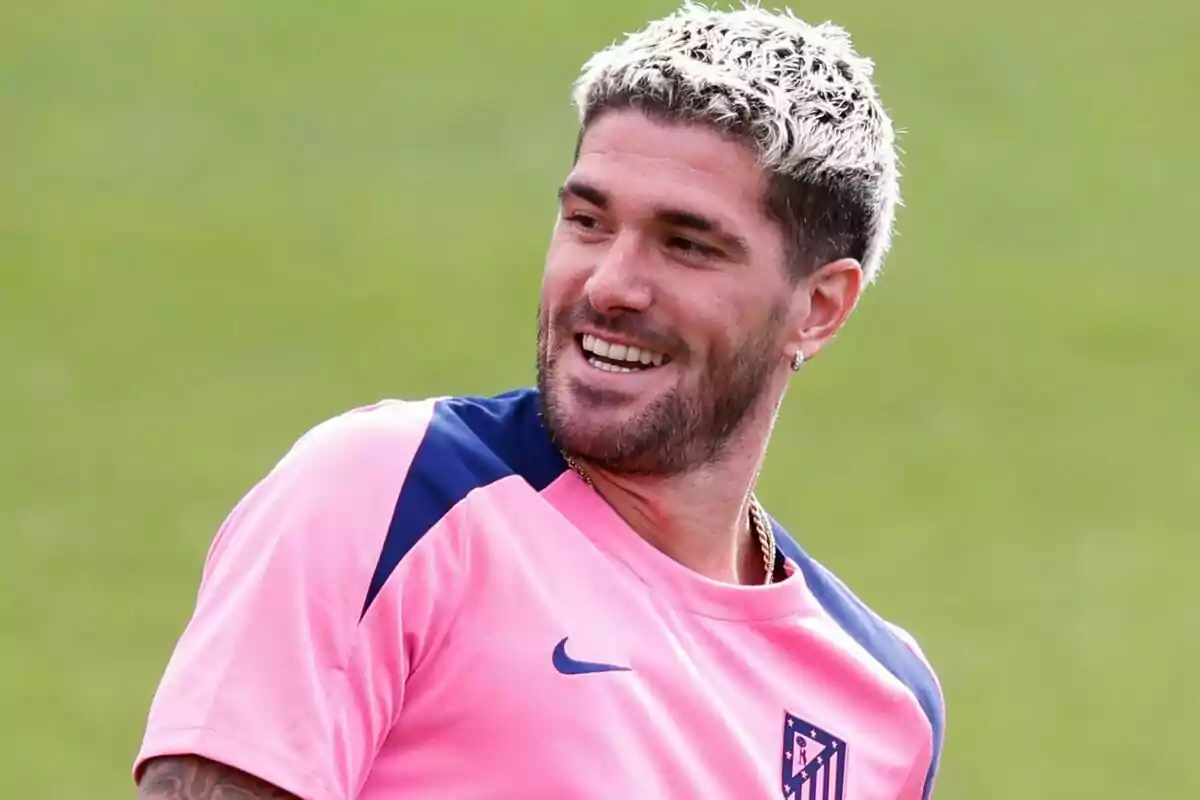 A man in a pink t-shirt and dyed blonde hair smiles as he looks to the side.