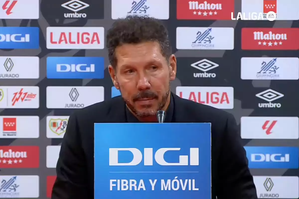 A bearded man with curly hair is speaking at a press conference, with a blue sign reading "DIGI FIBRA Y MÓVIL" in front of him and several sponsor logos in the background.