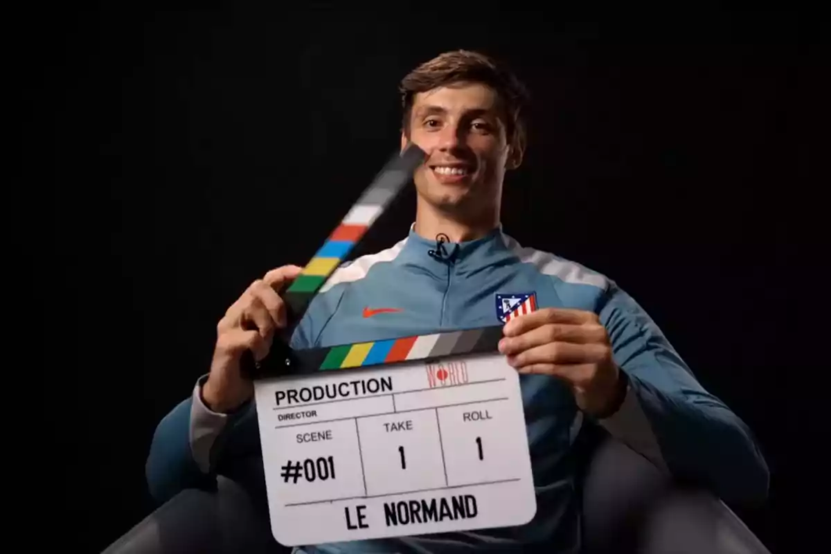 A smiling man in a blue sports jacket with a football team logo holds a film production clapperboard.