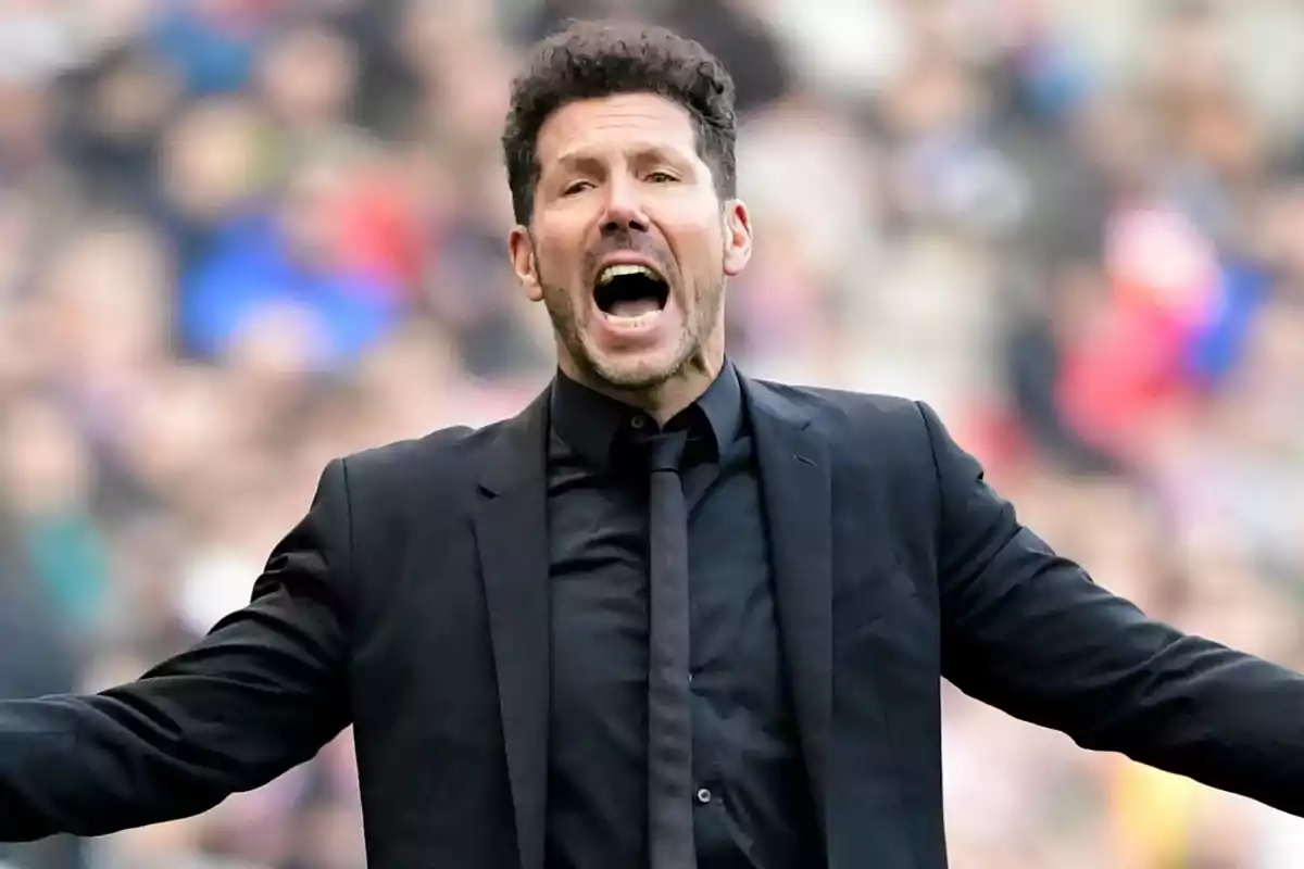 A man in a black suit gesturing in a sports environment.