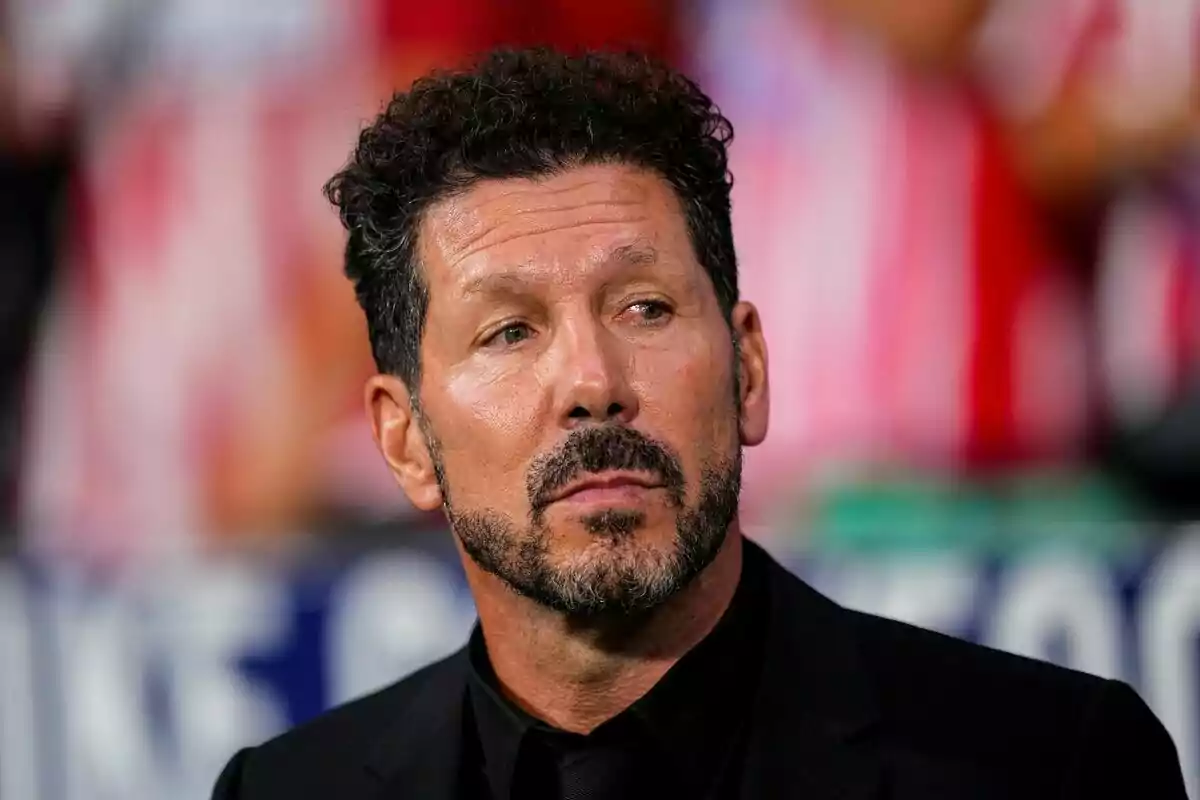 Man with beard and curly hair, dressed in a black suit, looking to the side with a serious expression.