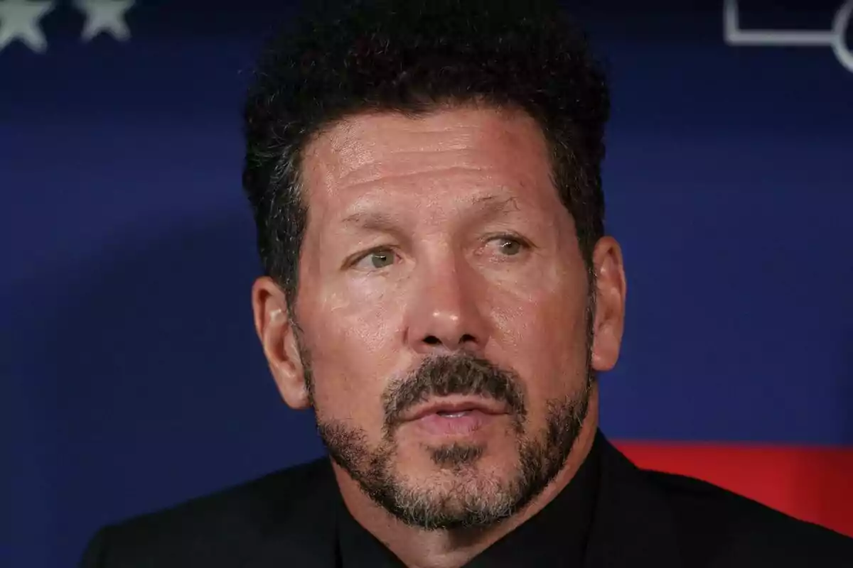 A man with a beard and curly hair looks to the side with a serious expression, against a dark blue background.