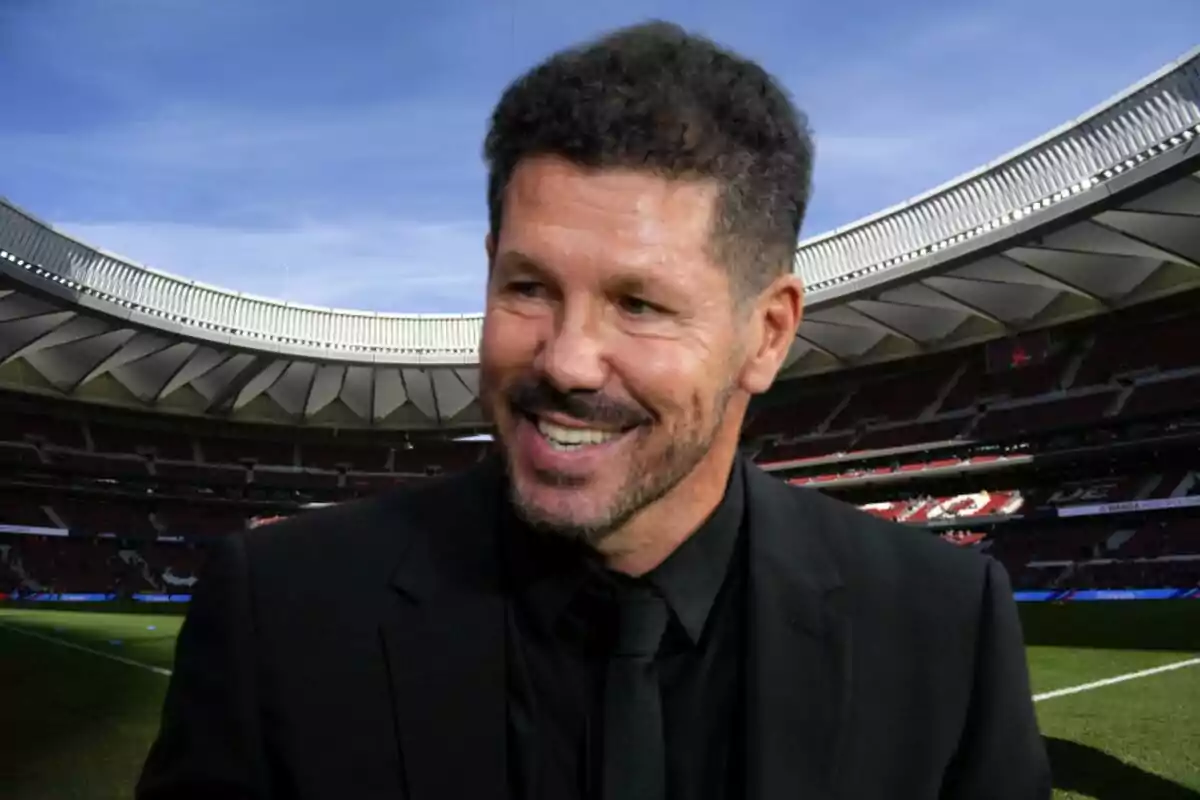 A smiling man with a beard and short hair, dressed in a black suit, in a football stadium.