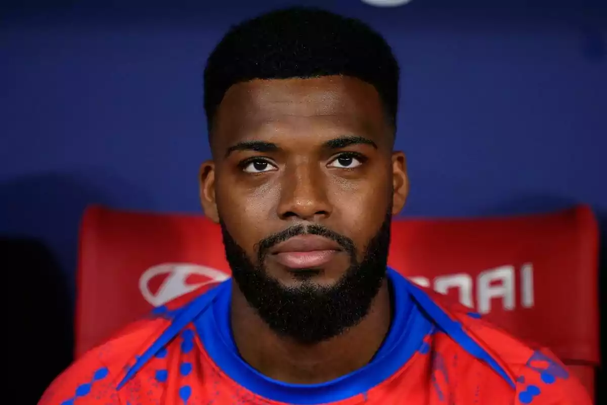 A man with a beard and short hair, wearing a red t-shirt with blue details, sitting in front of a dark background.