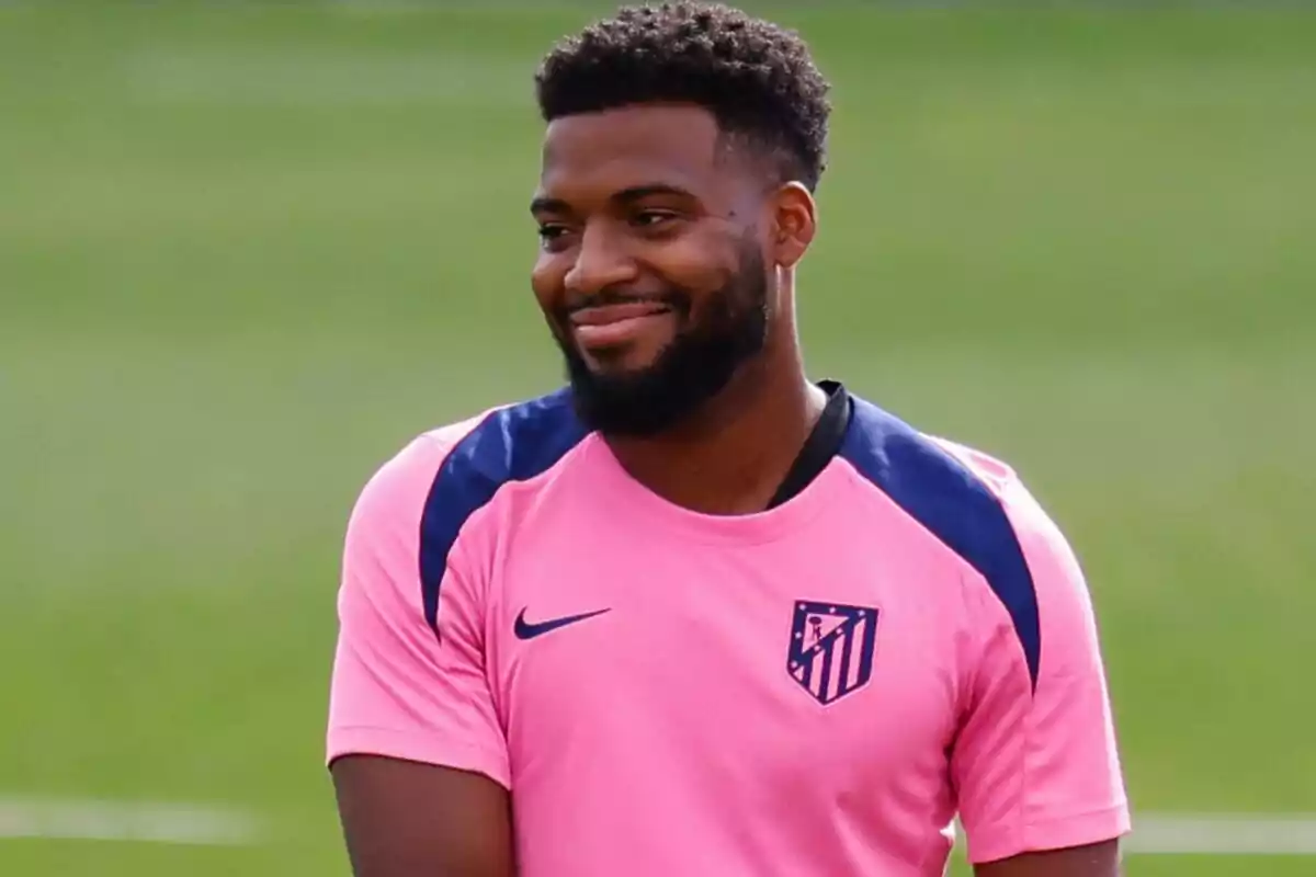 A soccer player wearing a pink Atletico Madrid jersey smiles at a training ground.