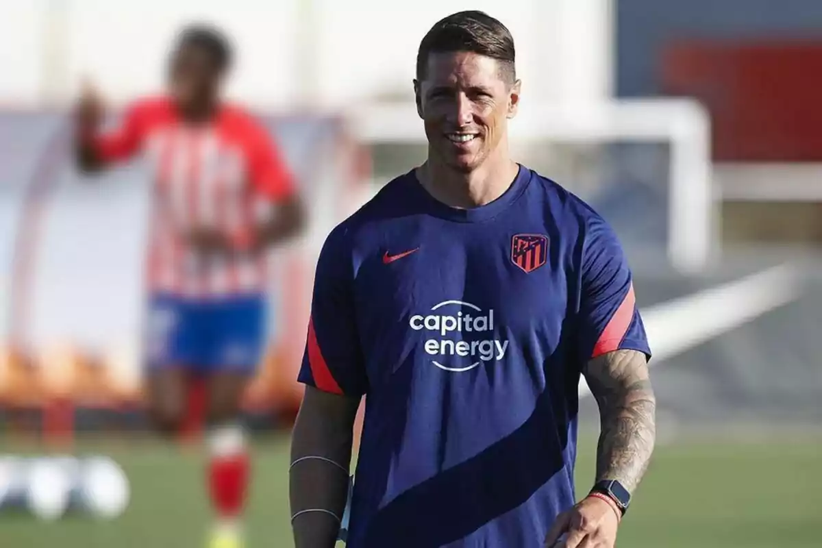 A man wearing an Atletico Madrid training shirt smiles as he walks on a football pitch.