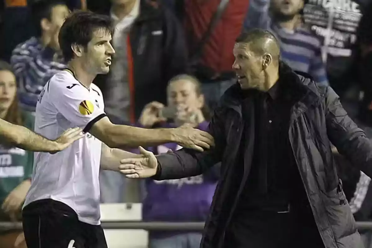 Two men greet each other on a football field, one of them wearing a white T-shirt and the other a dark coat.