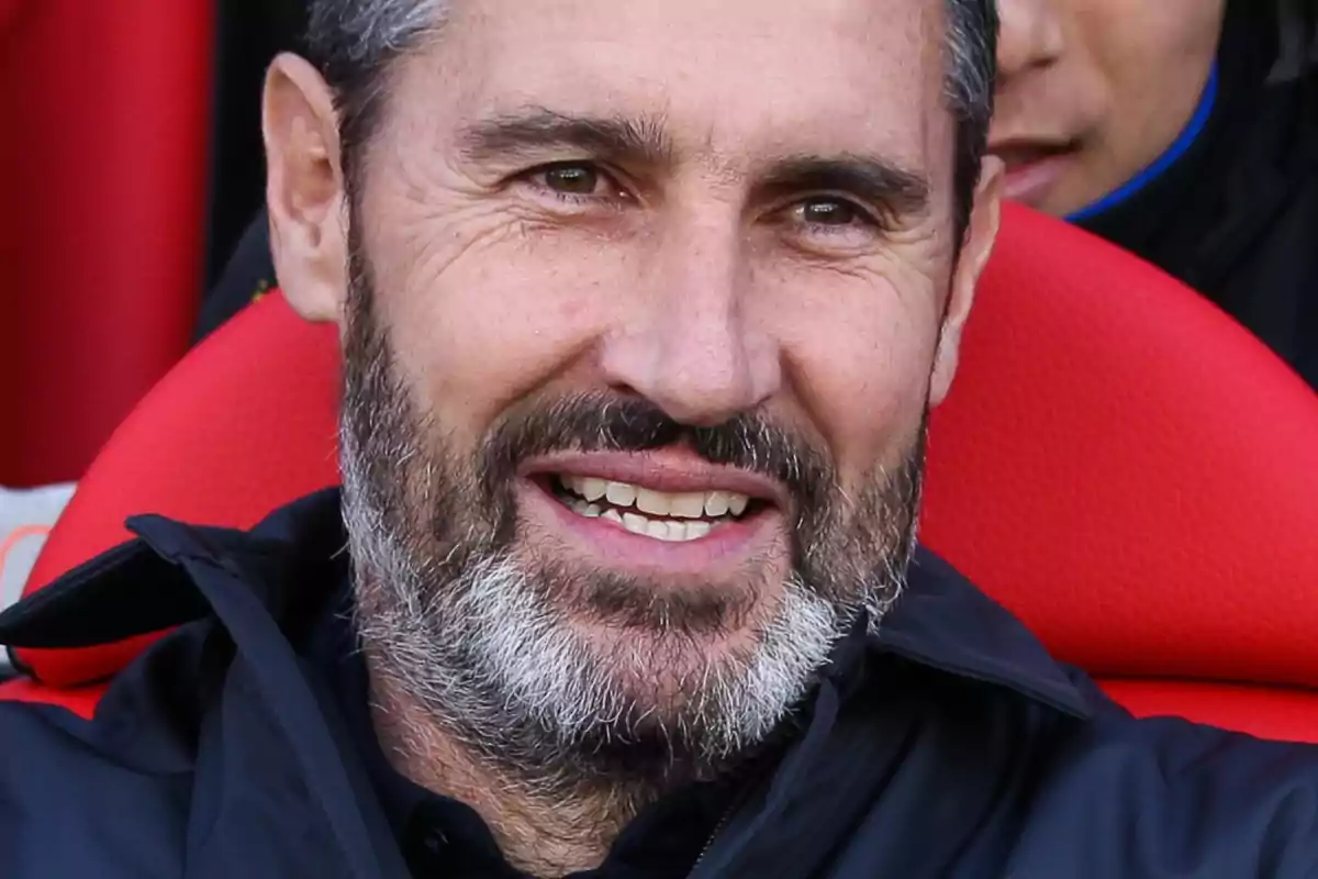 A man with a beard and short hair smiles while sitting on a red seat.