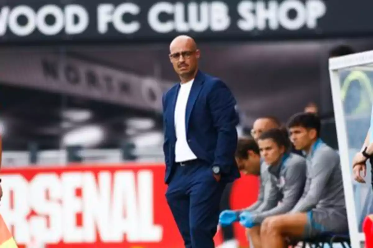 A football manager standing by the pitch, with players sitting on the bench behind him and an "Arsenal" sign in the background.