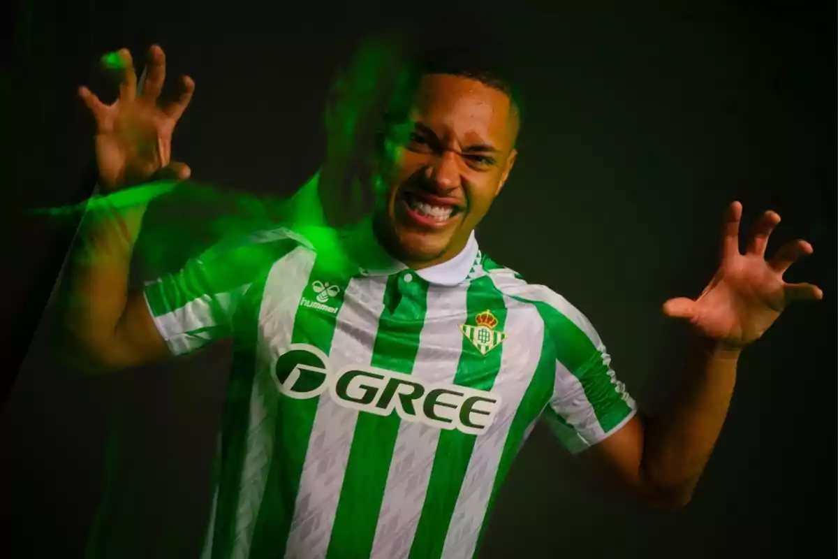 A football player wearing a Real Betis shirt makes an energetic gesture with his hands while smiling.