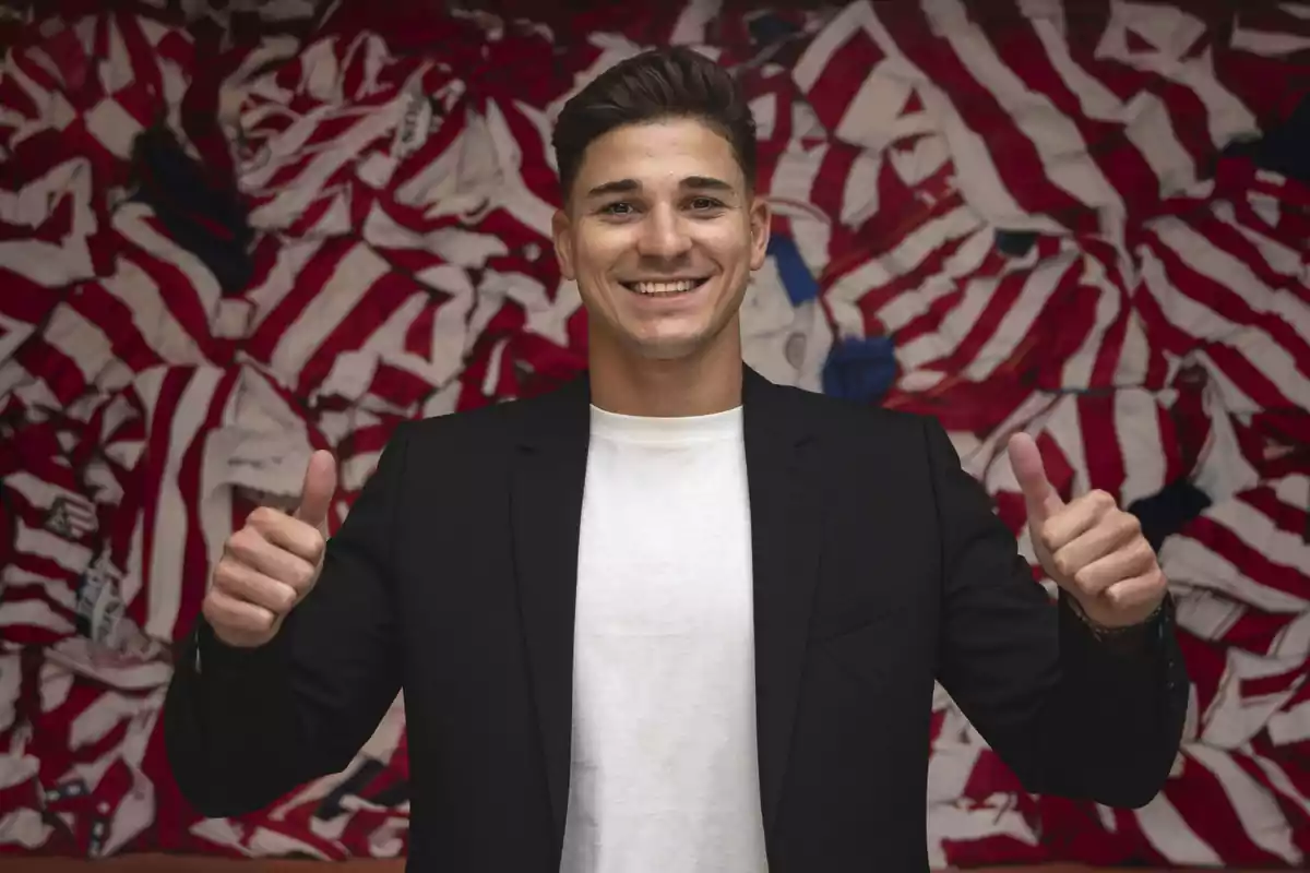 A smiling man in a white t-shirt and black jacket, giving both thumbs up in front of a background of red and white striped t-shirts.